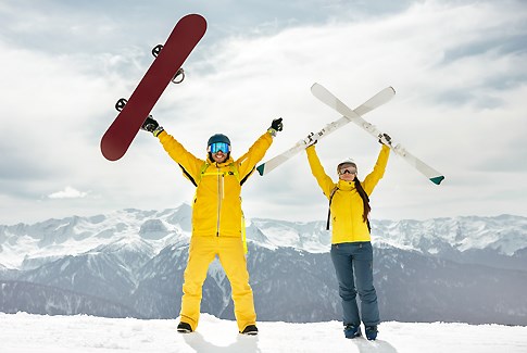 To skiløbere i gult løfter deres ski og snowboards op i luften