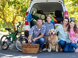 En familie med to børn og en hund sidder foran en åben varebil pakket til ferie, med cykel og picnickurv, omgivet af grøn natur og solskin.