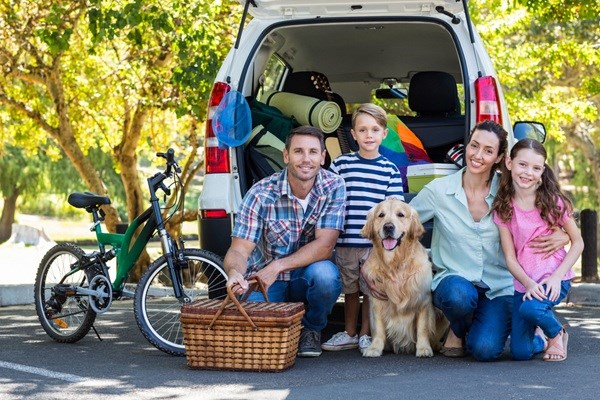 En familie med to børn og en hund sidder foran en åben varebil pakket til ferie, med cykel og picnickurv, omgivet af grøn natur og solskin.